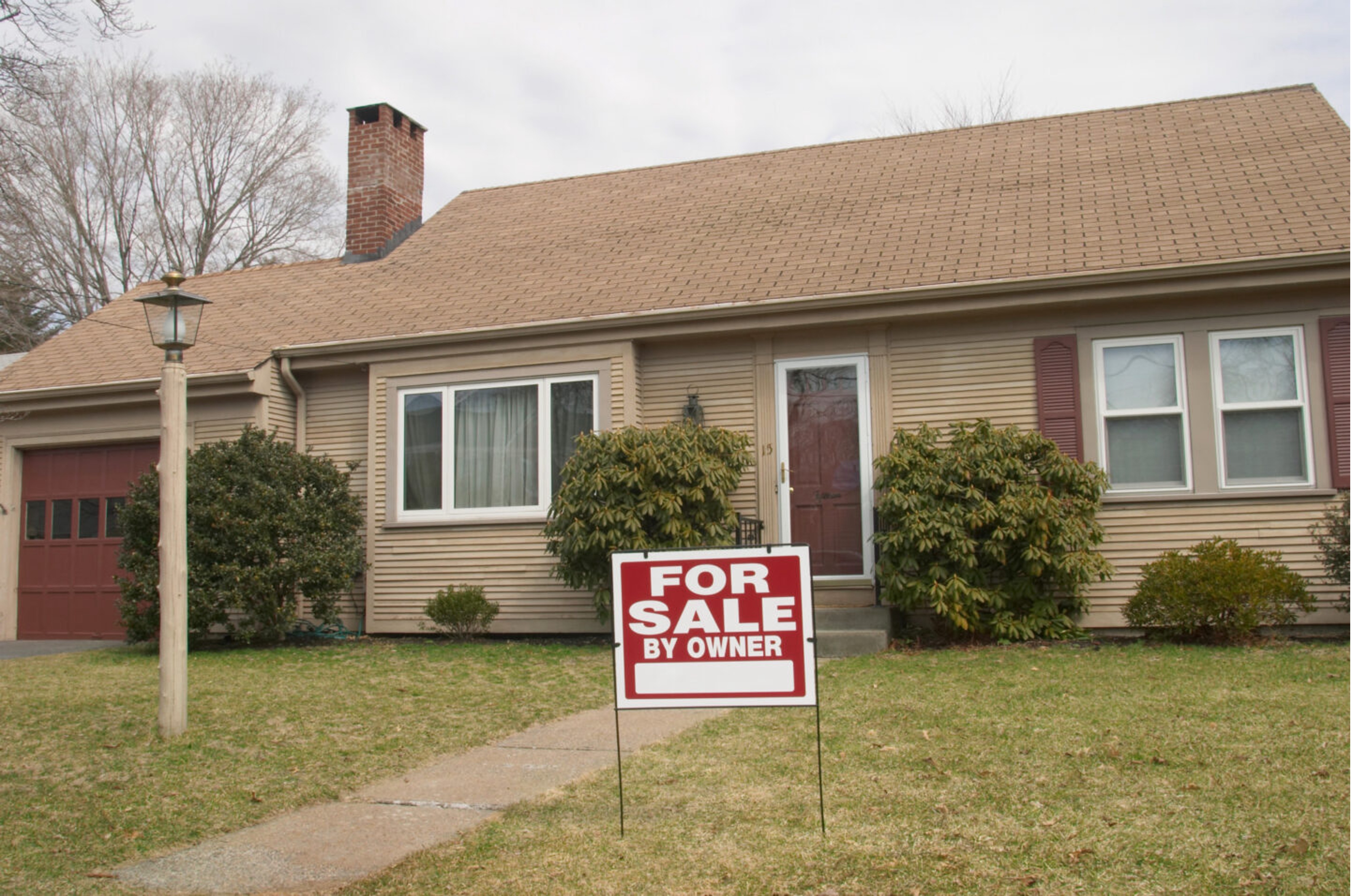 A one-story house with tan siding and a steep roof sits behind a "For Sale By Owner" sign in the front yard. Bushes frame the windows, creating a cozy feel.
